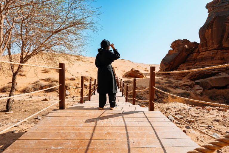 A guest on a wooden boardwalk between dunes and rocks in AlUla